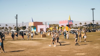 A lively outdoor festival scene with people walking and gathering in an open field. Colorful geometric structures with artistic designs are prominently displayed in the background amidst palm trees. The atmosphere is bright and sunny, indicative of a warm day.