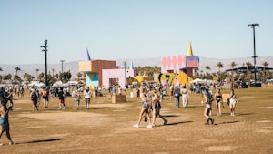 A lively outdoor festival scene with people walking and gathering in an open field. Colorful geometric structures with artistic designs are prominently displayed in the background amidst palm trees. The atmosphere is bright and sunny, indicative of a warm day.