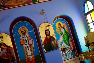 The image depicts several religious icons inside a church. The icons are painted in a classic style, featuring figures adorned with elaborate garments and halos. One icon shows a figure with a crown and ornate vestments holding a book, while another depicts a winged figure with a scroll. The background includes arched frames with rich details and colors, and there are stained glass windows visible on the side, adding a vibrant, colorful ambiance to the scene.