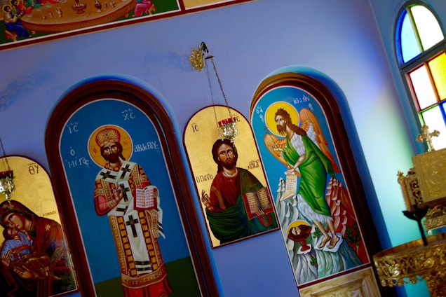 The image depicts several religious icons inside a church. The icons are painted in a classic style, featuring figures adorned with elaborate garments and halos. One icon shows a figure with a crown and ornate vestments holding a book, while another depicts a winged figure with a scroll. The background includes arched frames with rich details and colors, and there are stained glass windows visible on the side, adding a vibrant, colorful ambiance to the scene.