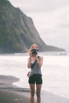 A person standing on a beach, capturing a photograph with a camera. The scene includes a large cliff or rock formation in the background. The atmosphere is overcast, with soft lighting creating a calming and serene environment.