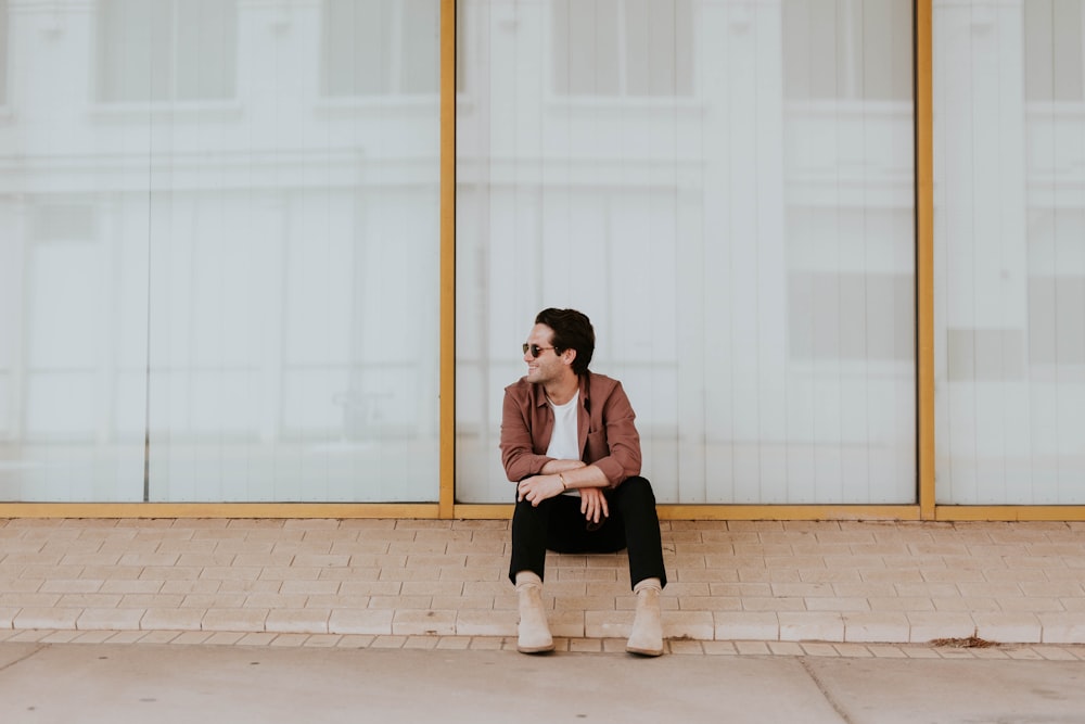 photo of man sitting on brown bricks against glass wall