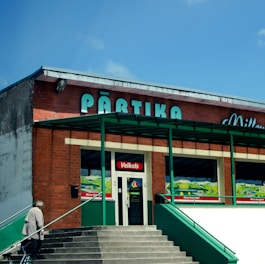 A brick building with green accents and a sign that reads 'Pārtika' on top. There is an entrance labeled 'Veikals' with stairs leading to it. A person is walking up the stairs carrying a bag.