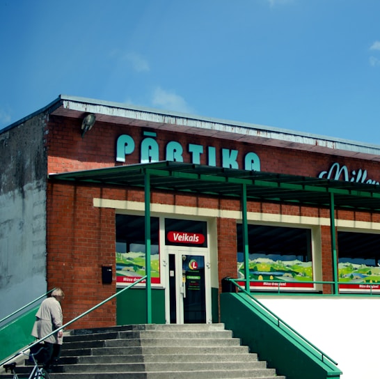 A brick building with green accents and a sign that reads 'Pārtika' on top. There is an entrance labeled 'Veikals' with stairs leading to it. A person is walking up the stairs carrying a bag.