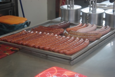 A variety of sausages are arranged on a metal tray, presumably for cooking or serving. The sausages differ in color and texture, indicating different types or flavors. In the background, there are stainless steel containers used for holding condiments or toppings.