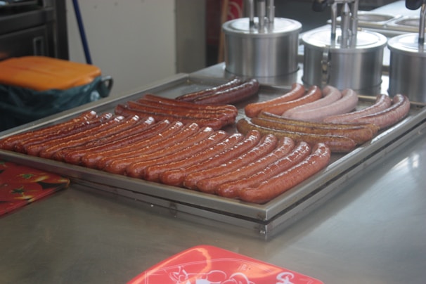 A variety of sausages are arranged on a metal tray, presumably for cooking or serving. The sausages differ in color and texture, indicating different types or flavors. In the background, there are stainless steel containers used for holding condiments or toppings.