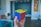 A hand holding a solved Rubik's Cube with red, blue, and yellow faces visible. The background shows a modern interior with colorful chairs and a wooden decorative piece on the wall.