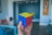 A hand holding a solved Rubik's Cube with red, blue, and yellow faces visible. The background shows a modern interior with colorful chairs and a wooden decorative piece on the wall.