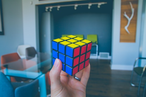 A hand holding a solved Rubik's Cube with red, blue, and yellow faces visible. The background shows a modern interior with colorful chairs and a wooden decorative piece on the wall.
