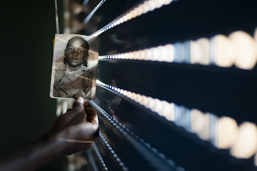 A hand holds a black and white photograph of a child against a backdrop of horizontal light slats creating a pattern of light and shadow. The photograph is slightly angled, and the light casts a warm glow. The scene conveys a sense of nostalgia and contemplation.