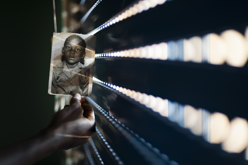 A hand holds a black and white photograph of a child against a backdrop of horizontal light slats creating a pattern of light and shadow. The photograph is slightly angled, and the light casts a warm glow. The scene conveys a sense of nostalgia and contemplation.