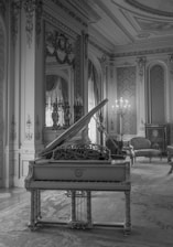 A luxurious and ornately decorated room features a grand piano with intricate designs. The room is adorned with elaborate moldings, a high ceiling, and large windows with heavy drapery. Elegant furniture, including a sofa and candelabra, adds to the opulence of the setting.