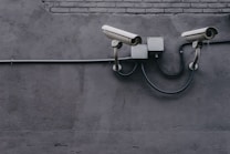 Two security cameras are mounted on a gray concrete wall, connected by a black cable. The wall has a rough texture, and there is a segment of brickwork near the top.