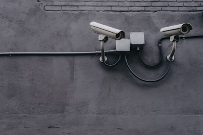 Two security cameras are mounted on a gray concrete wall, connected by a black cable. The wall has a rough texture, and there is a segment of brickwork near the top.