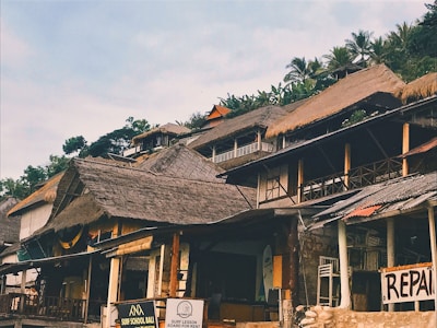 A collection of thatched-roof buildings on a hillside with lush greenery in the background. Signs indicate a surf school and a repair shop, suggesting a coastal or tropical setting.