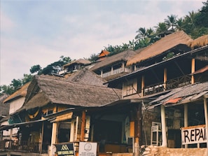 A collection of thatched-roof buildings on a hillside with lush greenery in the background. Signs indicate a surf school and a repair shop, suggesting a coastal or tropical setting.