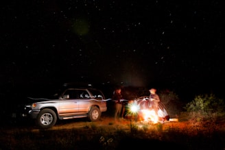 Nighttime camp setup with a glowing campfire beside a parked SUV under a star-filled sky.