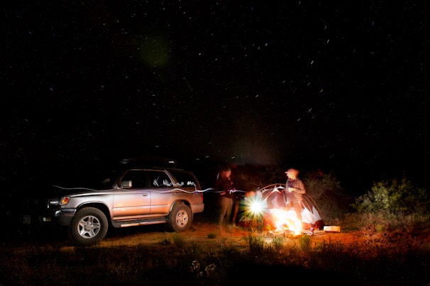 Nighttime camp setup with a glowing campfire beside a parked SUV under a star-filled sky.