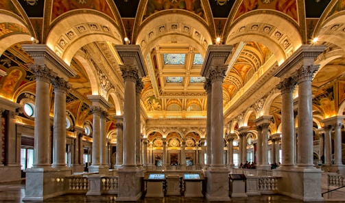 An opulent interior hall with ornate architecture featuring grand arches, marble columns, and elaborately decorated ceilings. Intricate patterns and frescoes adorn the ceiling, with warm lighting enhancing the luxurious ambiance. The space is open and spacious, with a classic and historical feel.