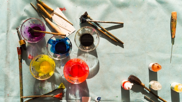 A variety of paintbrushes and open jars of paint in vibrant colors such as purple, blue, black, yellow, and red are spread across a light-colored surface. The brushes appear to have been used, with traces of paint on their bristles. Other painting tools and small paint bottles are scattered around, suggesting an artistic workspace.