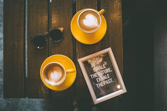 Two yellow cups of coffee with latte art sit on a wooden table next to a square-framed motivational quote that reads 'INHALE THE FUTURE, EXHALE THE PAST.' A pair of black sunglasses rests near the cups.