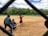 A group of people engaged in a casual game of baseball on a public field. In the foreground, a player wearing a red jersey has just swung at a pitch. Two other players, one acting as the catcher and another as the umpire, are crouched behind him. In the background, players are scattered across the field, which is surrounded by lush green trees under a partly cloudy sky.