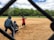 A group of people engaged in a casual game of baseball on a public field. In the foreground, a player wearing a red jersey has just swung at a pitch. Two other players, one acting as the catcher and another as the umpire, are crouched behind him. In the background, players are scattered across the field, which is surrounded by lush green trees under a partly cloudy sky.