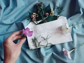 A hand holding a pink flower is placed on light blue fabric. An envelope decorated with flowers contains handwritten text in Cyrillic, surrounded by various fresh flowers and greenery.