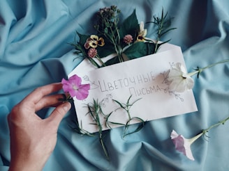 A hand holding a pink flower is placed on light blue fabric. An envelope decorated with flowers contains handwritten text in Cyrillic, surrounded by various fresh flowers and greenery.