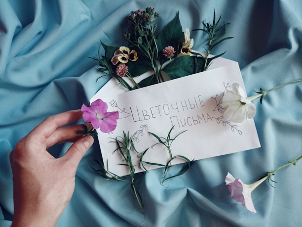 A hand holding a pink flower is placed on light blue fabric. An envelope decorated with flowers contains handwritten text in Cyrillic, surrounded by various fresh flowers and greenery.