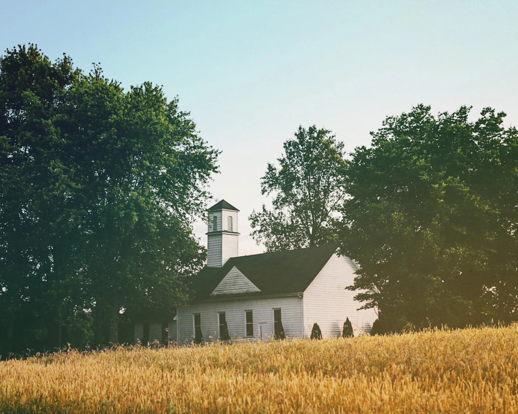 A white colonial-era church building with a modest bell tower surrounded by mature green trees