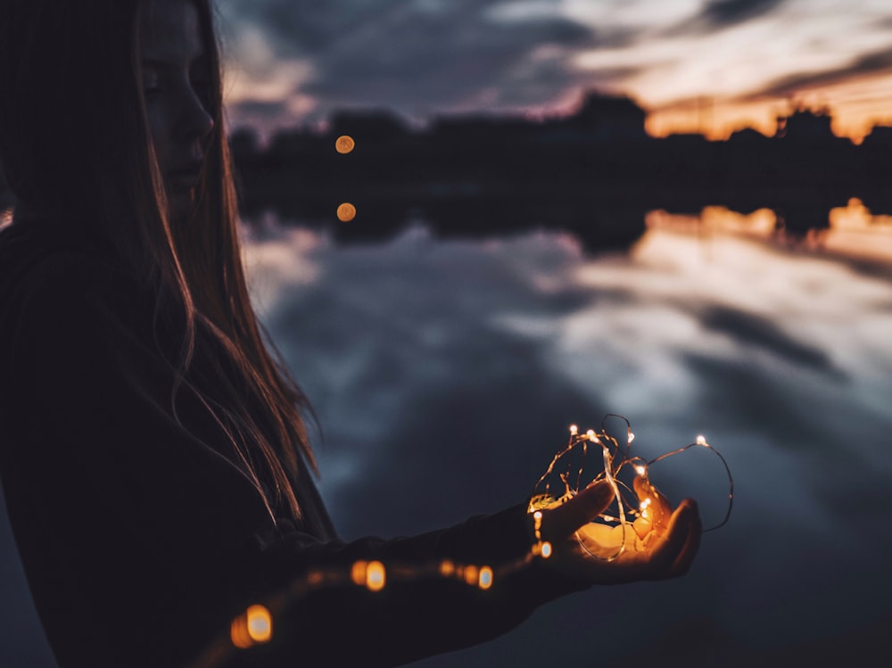 A girl holding fairy lights in her hand by a lake on an evening