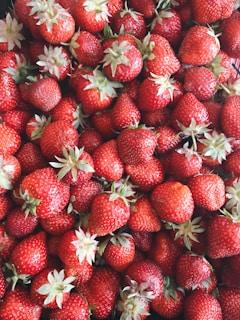 A large pile of fresh strawberries is tightly packed together with some showing their green leaves and stems. The strawberries appear ripe, vibrant, and ready to eat.