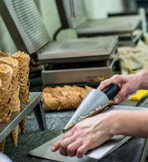 A person is manually rolling waffle cones on a metal surface. Several freshly made cones are arranged in stacks nearby. The setting appears to be a kitchen or a production area, with professional equipment designed for making waffle cones.