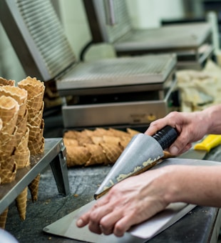 A person is manually rolling waffle cones on a metal surface. Several freshly made cones are arranged in stacks nearby. The setting appears to be a kitchen or a production area, with professional equipment designed for making waffle cones.