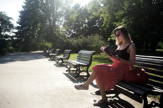 A young woman sitting on a park bench, reading a book with soft pink tones around her.