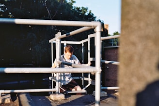 A young person is sitting with a pensive expression on an elevated platform surrounded by metal railings. The background includes lush green trees and part of a building, with sunlight casting shadows on the scene.
