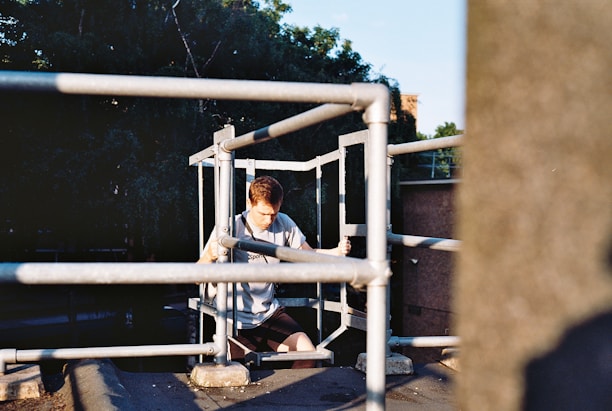 A young person is sitting with a pensive expression on an elevated platform surrounded by metal railings. The background includes lush green trees and part of a building, with sunlight casting shadows on the scene.