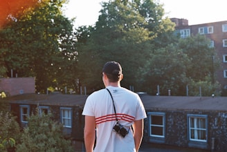 A person wearing a white shirt and cap is standing on a rooftop, holding a camera. The background features lush green trees and several residential buildings under a clear sky.