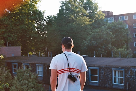 A person wearing a white shirt and cap is standing on a rooftop, holding a camera. The background features lush green trees and several residential buildings under a clear sky.