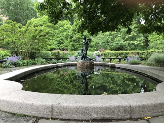 A tranquil garden scene featuring a round stone-edged pond with a bronze statue in the center. Surrounding the pond are lush green trees and shrubs with purple and pink flowers. The water reflects the greenery and statue, enhancing the peaceful ambiance.