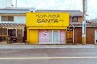 A small, yellow building labeled 'ペットハウス SANTA' stands alongside other buildings with traditional architecture. The structure appears to be a pet store with a dog silhouette in the window display. Several plants are placed near the entrance.