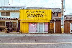 A small, yellow building labeled 'ペットハウス SANTA' stands alongside other buildings with traditional architecture. The structure appears to be a pet store with a dog silhouette in the window display. Several plants are placed near the entrance.