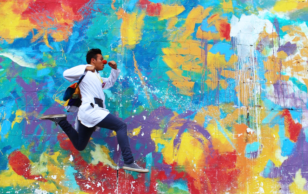 A dark-skinned male leaping along the sidewalk in front of an abstractly painted wall.