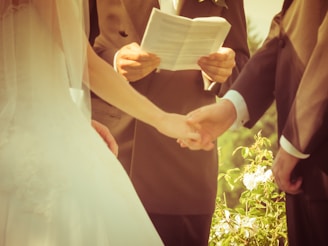A close-up view of two people holding hands during a wedding ceremony. There is an officiant in the background holding open a book. The setting has a warm tone and the background features greenery and flowers.