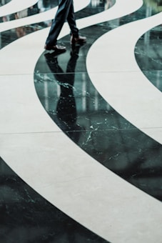 Curved, flowing patterns on a polished floor with a reflection of a person's legs wearing formal trousers and shoes, creating an abstract visual effect.