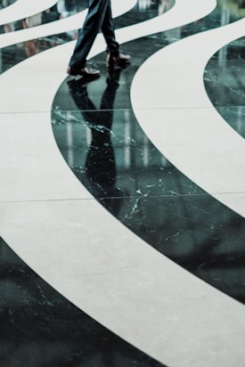 Curved, flowing patterns on a polished floor with a reflection of a person's legs wearing formal trousers and shoes, creating an abstract visual effect.