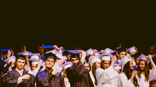 A group of graduates wearing caps and gowns are standing together during a ceremony. They are raising their hands to adjust their caps. Most of them are smiling, and the gowns vary in color between black and white.
