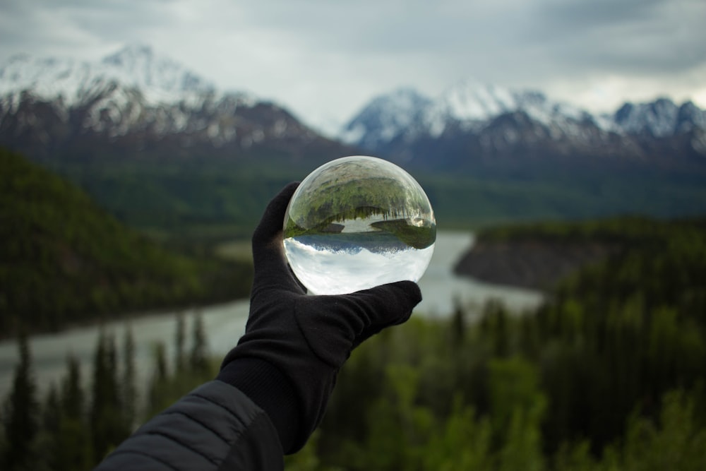 A person's hand in a glove holding a large glass ball against the backdrop of snowy mountains