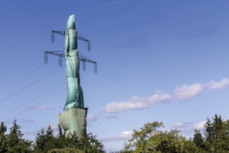 A high-voltage power transmission tower wrapped in green protective netting rises above lush green trees. The tower is set against a backdrop of a clear, blue sky with sparse clouds.